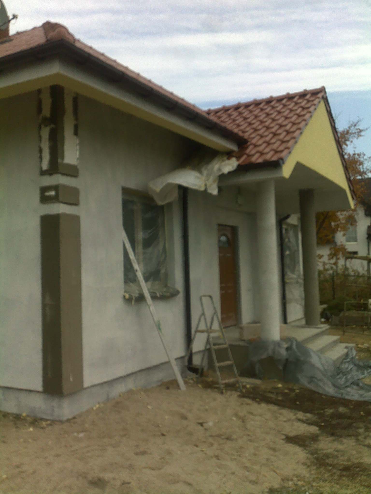 Dom jednorodzinny w trakcie prac elewacyjnych: ściany pokryte surowym tynkiem, widoczne profile narożne, okna zabezpieczone folią, drabina oparta o ścianę, teren wokół domu częściowo uprzątnięty.