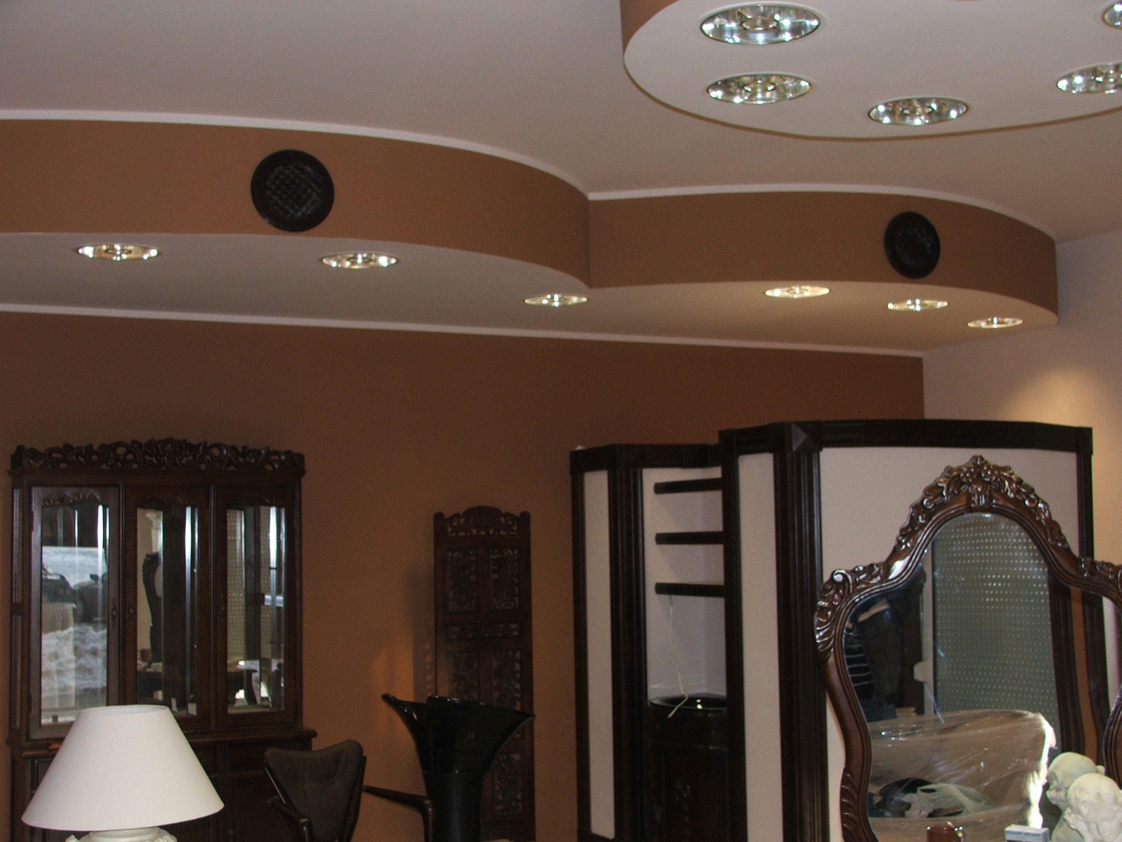 Sufit z gładzią gipsową, okrągłymi wnękami z oświetleniem punktowym i kratkami wentylacyjnymi, w tle meble w stylu retro.