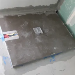 Przygotowanie posadzki w łazience pod płytki: wylewka samopoziomująca, hydroizolacja w narożnikach, zabezpieczone rury, widoczna taśma malarska i wałek.