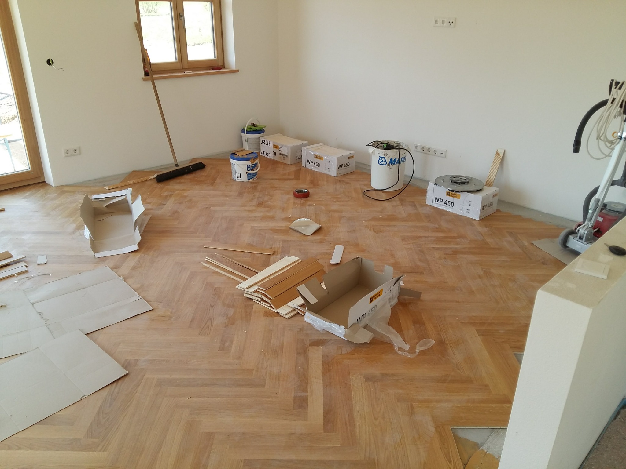 Układanie parkietu w jodełkę: widoczne puste ściany, okno, materiały budowlane, kartony, maszyna do polerowania i szczotka w trakcie prac wykończeniowych.