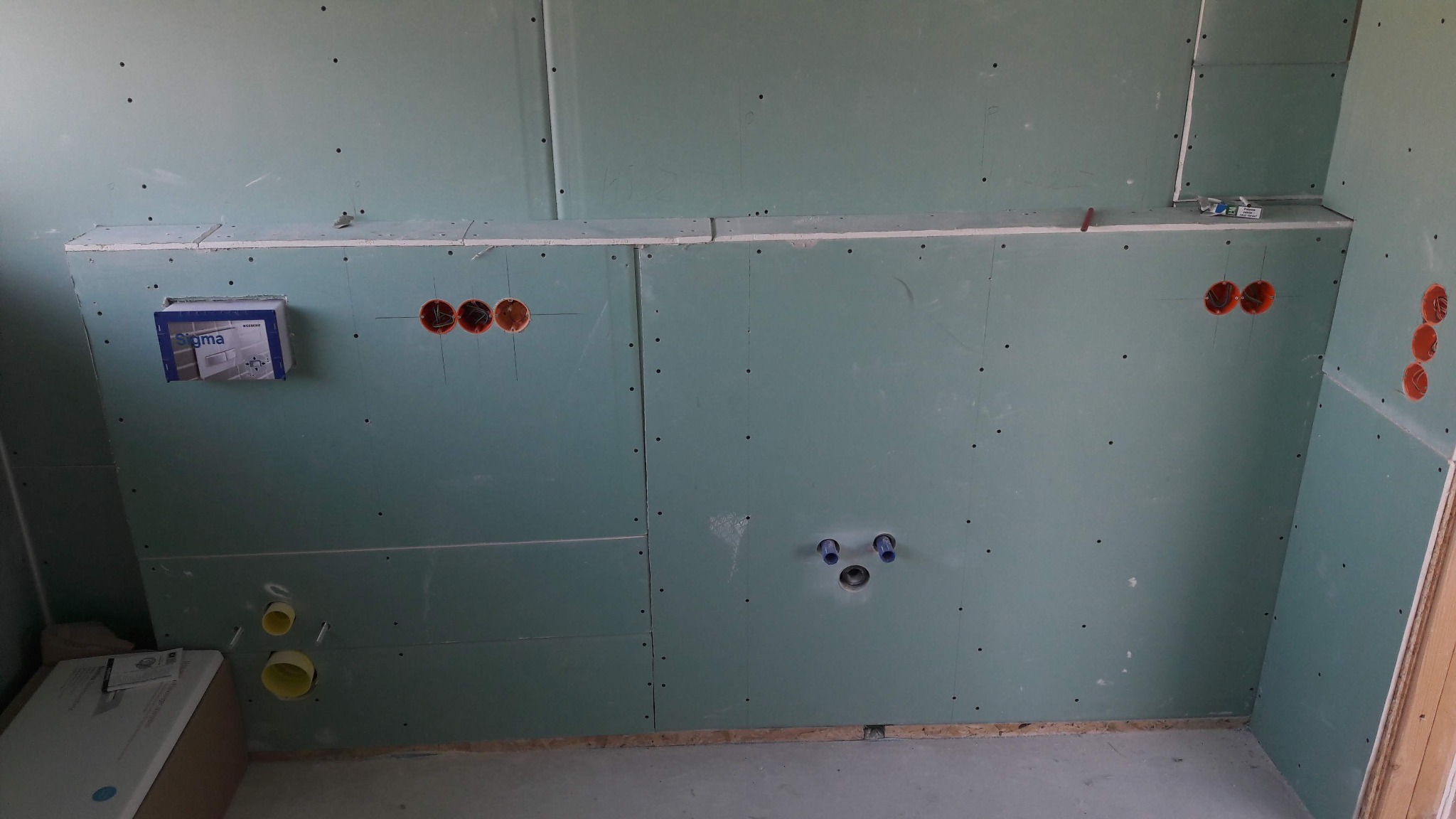 Ściana w trakcie przygotowania do wykończenia w łazience, widoczne płyty gipsowo-kartonowe, instalacja wodno-kanalizacyjna i elektryczna w puszkach, przygotowane podłączenie do umywalki i baterii.