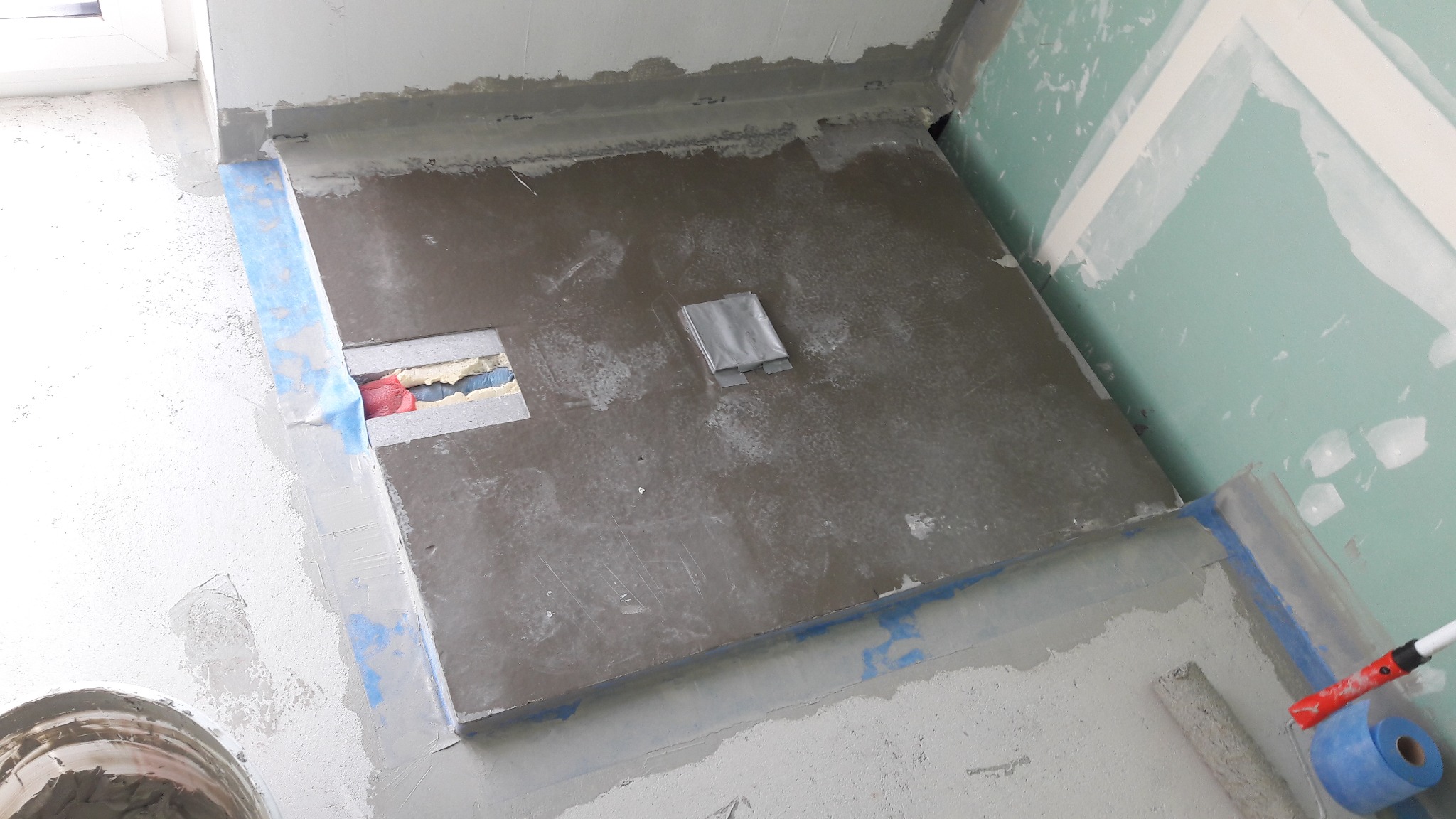 Przygotowanie posadzki w łazience pod płytki: wylewka samopoziomująca, hydroizolacja w narożnikach, zabezpieczone rury, widoczna taśma malarska i wałek.