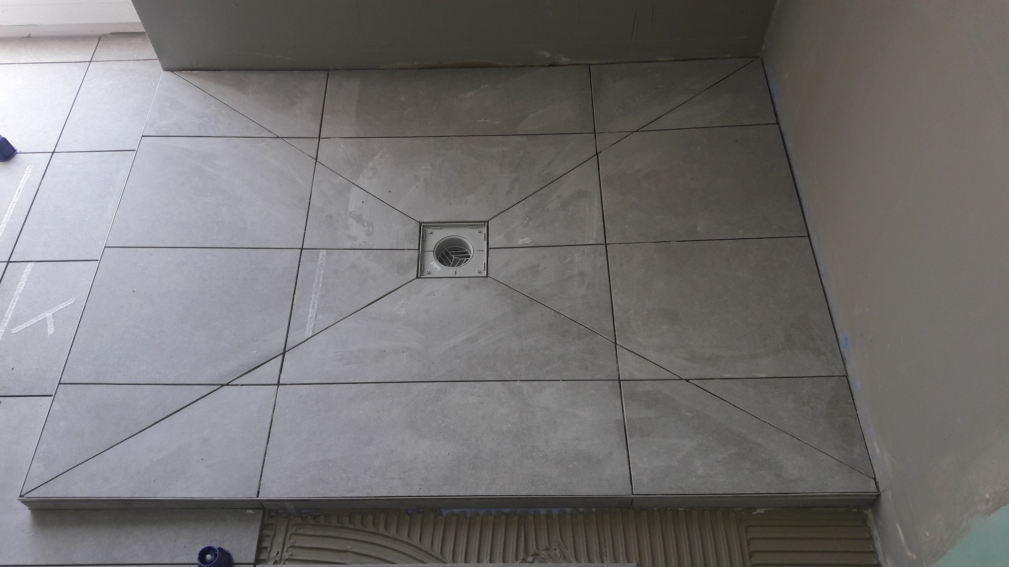 Szare płytki podłogowe ułożone w kwadraty, z centralnym odpływem liniowym, zaznaczone linie cięcia płytek.