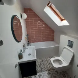 Nowoczesna łazienka na poddaszu ze skośnym sufitem, z umywalką na szafce, owalnym lustrem, wanną obudowaną r&oacute;żowymi płytkami, toaletą i wzorzystą podłogą.