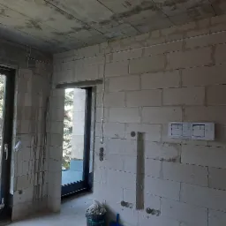 Surowy stan wnętrza w trakcie prac instalacyjnych: widoczne przewody elektryczne prowadzone po ścianie z betonu komórkowego, otwory pod gniazdka i włączniki, plany techniczne przyklejone do ściany,...