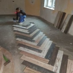 JT-KA M. I P. WĄSOWSCY SPÓŁKA JAWNA - Układanie drewnopodobnych paneli podłogowych w jodełkę w surowym wnętrzu. Widoczny pracownik z laserowym niwelatorem i wiadro kleju. Częściowo ułożona podłoga.
