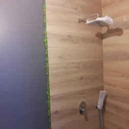 Fragment łazienki w Krakowie z wykończoną strefą prysznicową: szara ściana z płytek, drewnopodobne panele ścienne, pionowy pasek mchu dekoracyjnego, bateria i słuchawka prysznicowa w folii...