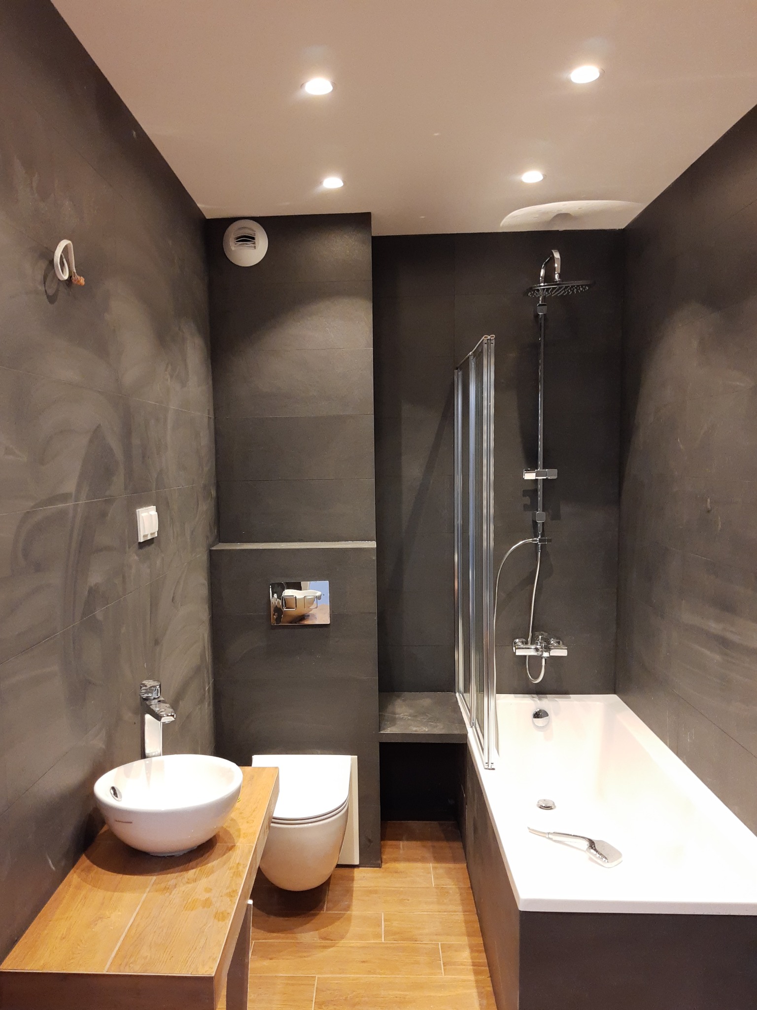 Nowoczesna łazienka z ciemnymi, strukturalnymi ścianami, drewnianym blatem pod umywalką, białą armaturą i kabiną prysznicową nad wanną.