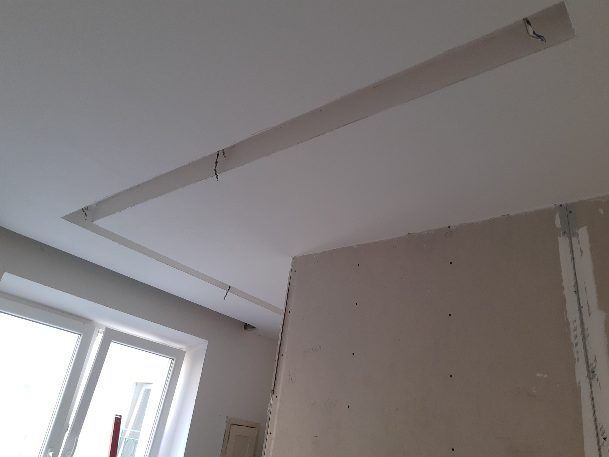Wnętrze w trakcie wykańczania: sufit z wnęką i wystającymi kablami, ściana z płyt kartonowo-gipsowych z widocznymi śrubami i notatkami, okno z widokiem na zewnątrz.