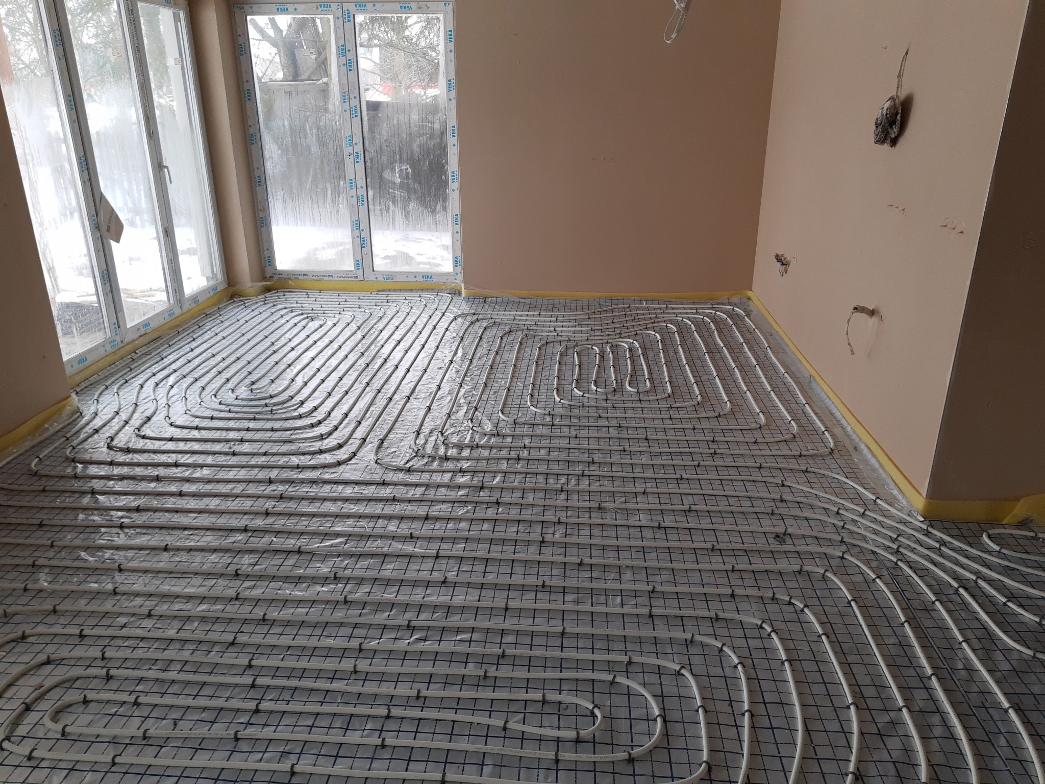 Rozłożone rury ogrzewania podłogowego na folii z siatką w jasnym pomieszczeniu z dużym oknem, widoczne wystające kable ze ściany.