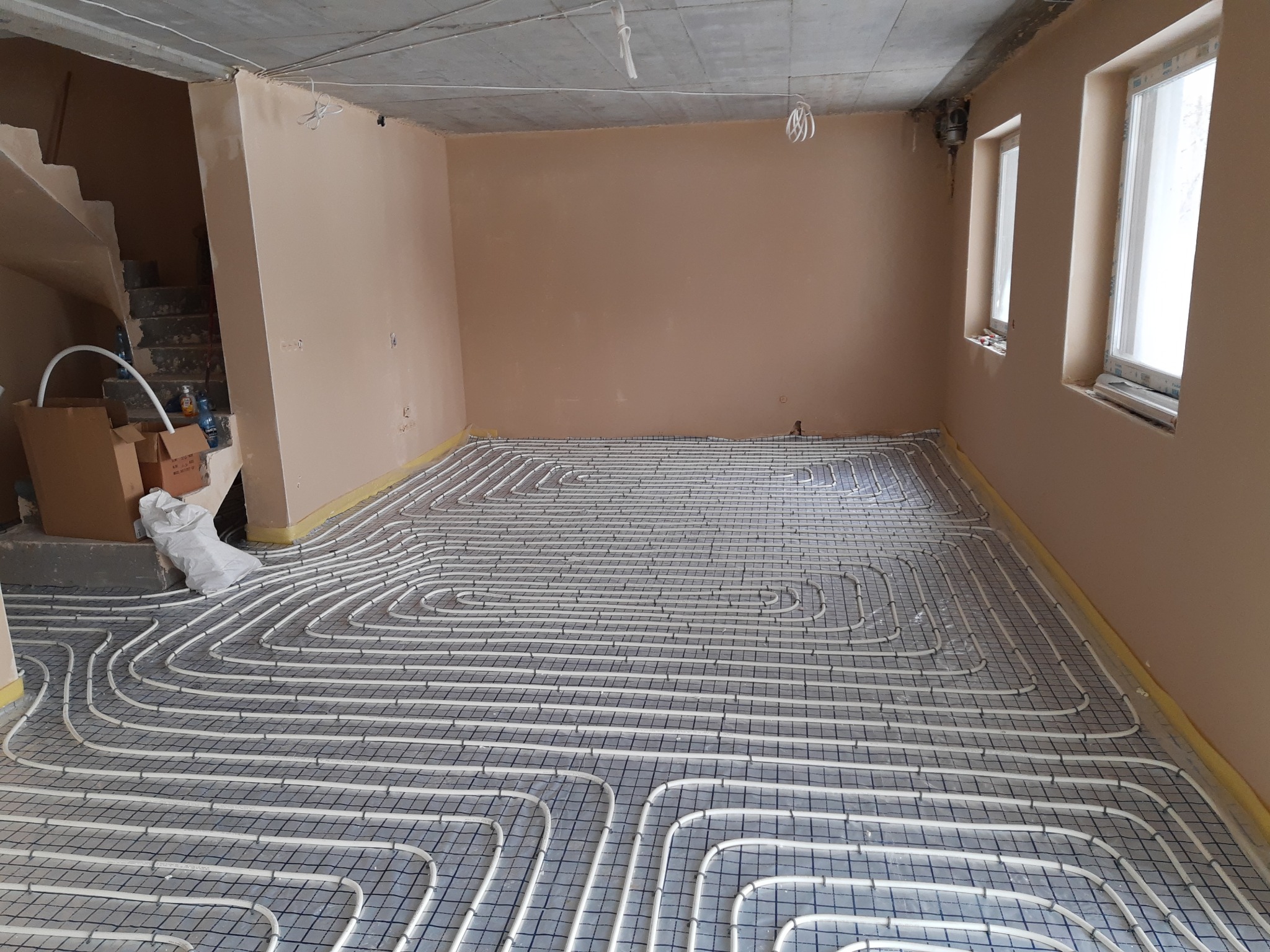 Rozłożona instalacja ogrzewania podłogowego z rur na siatce w surowym pomieszczeniu przed wylewką, widoczne ściany w kolorze beżowym i okna.
