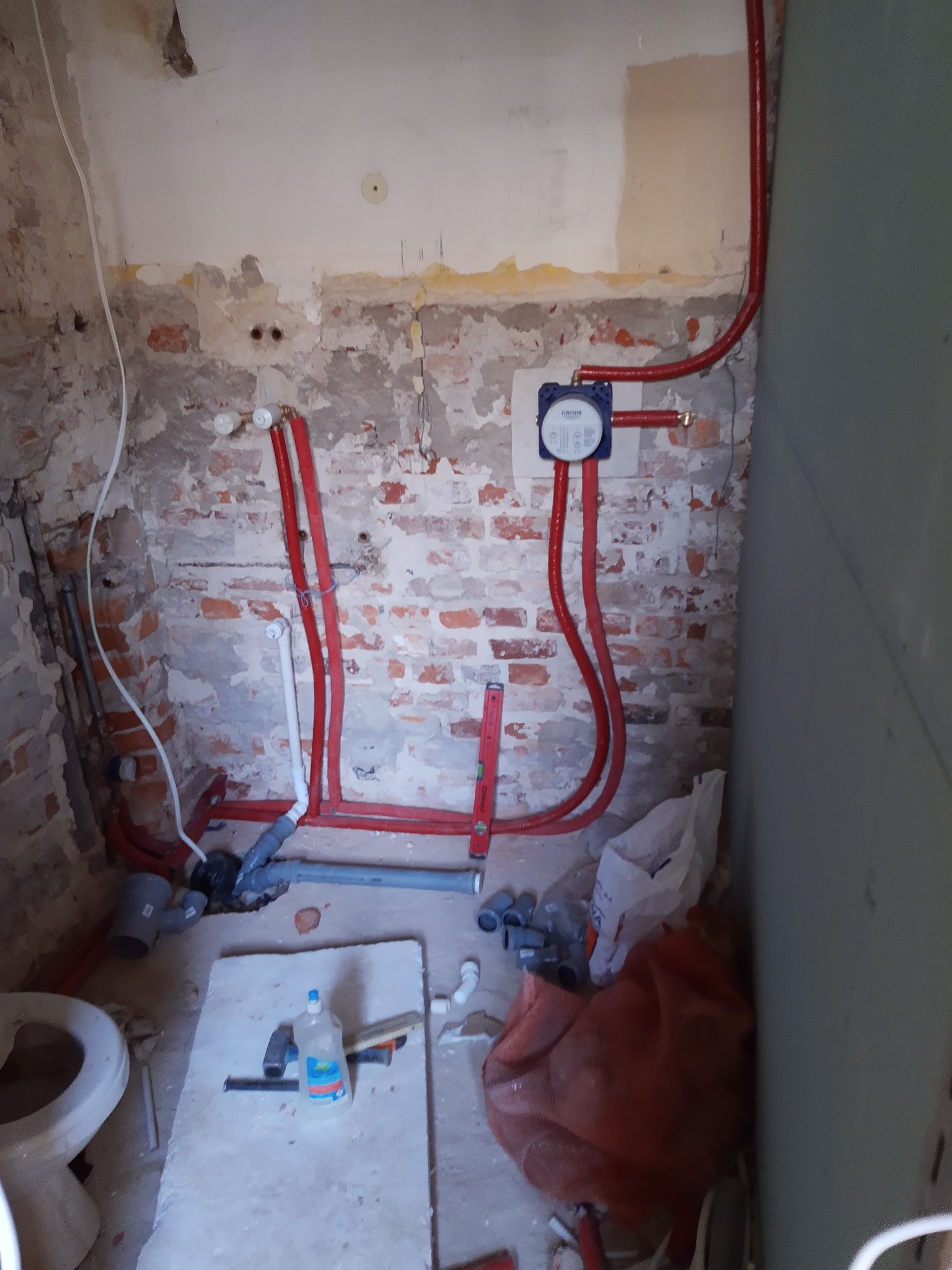 Surowy stan łazienki w trakcie prac: widoczne czerwone rury instalacji wodnej, nieotynkowana cegła, oraz elementy podłączenia toalety.