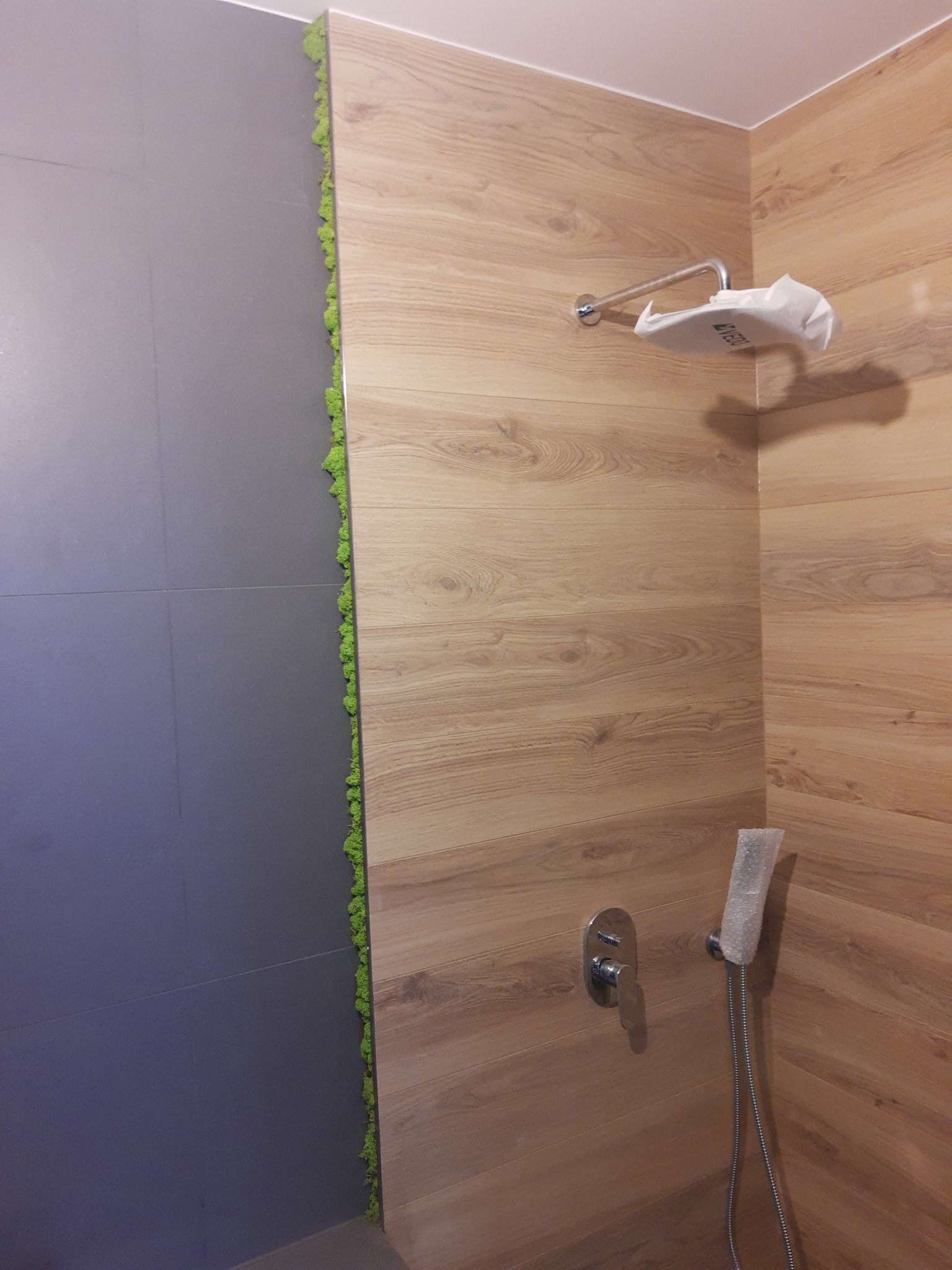 Fragment łazienki w Krakowie z wykończoną strefą prysznicową: szara ściana z płytek, drewnopodobne panele ścienne, pionowy pasek mchu dekoracyjnego, bateria i słuchawka prysznicowa w folii...