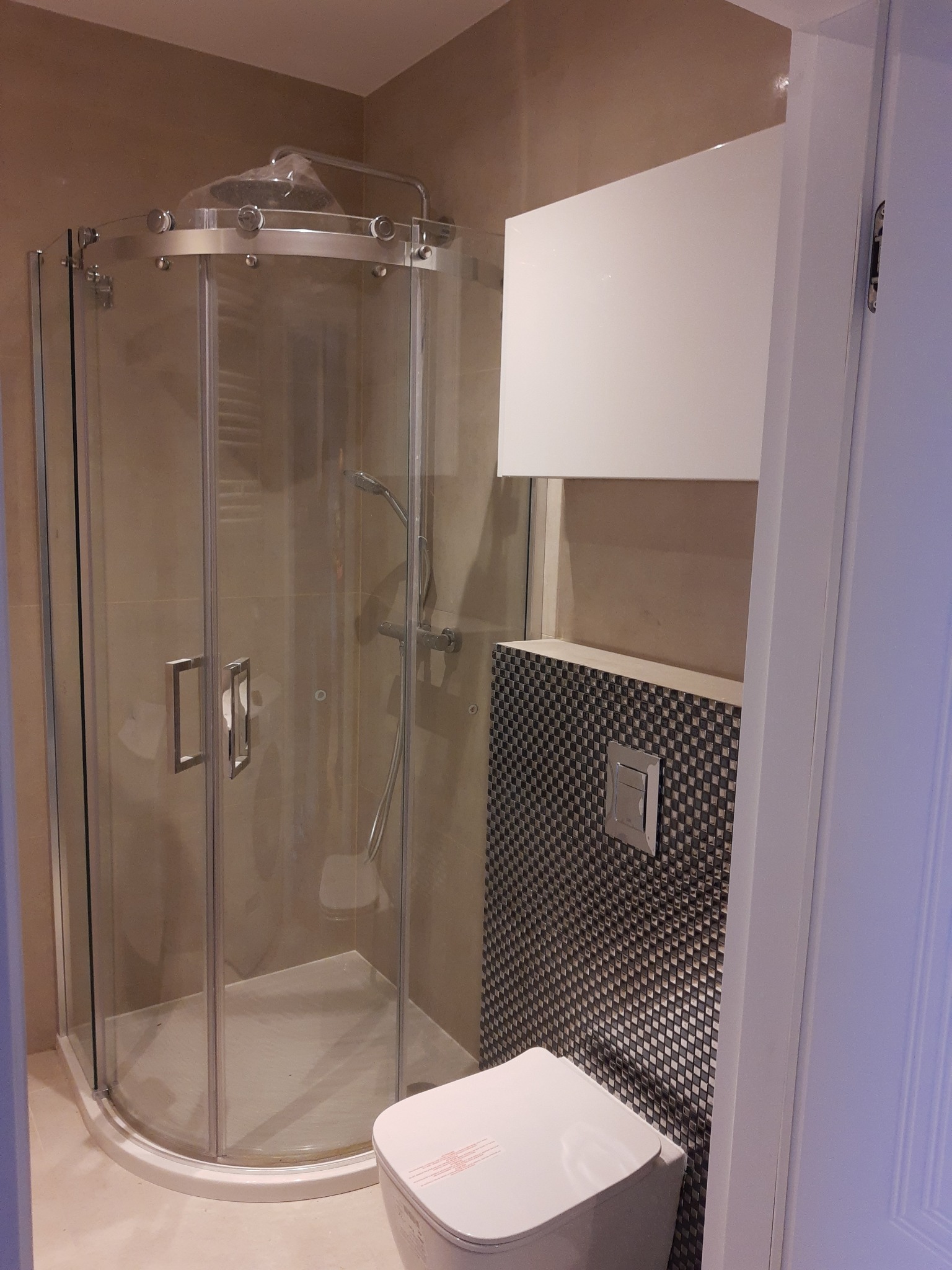 Narożna kabina prysznicowa z chromowanymi okuciami, wisząca toaleta z przyciskiem spłukującym na ścianie wyłożonej mozaiką w odcieniach szarości, biała szafka nad toaletą.