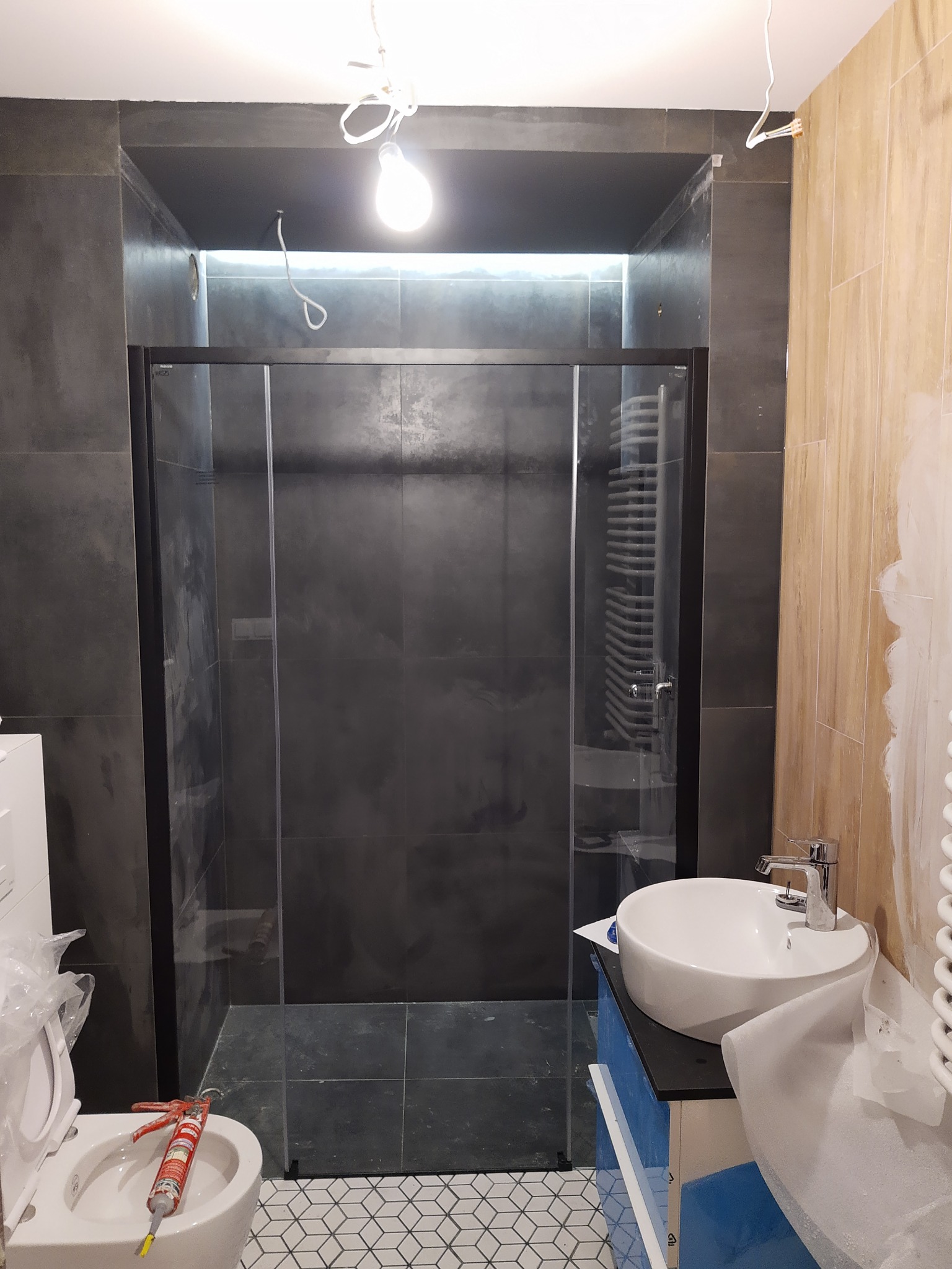 Ciemna łazienka w trakcie remontu: kabina prysznicowa z czarnymi profilami, umywalka nablatowa na niebieskiej szafce, biała toaleta z silikonem w tubie na desce, widoczne przewody oświetleniowe.