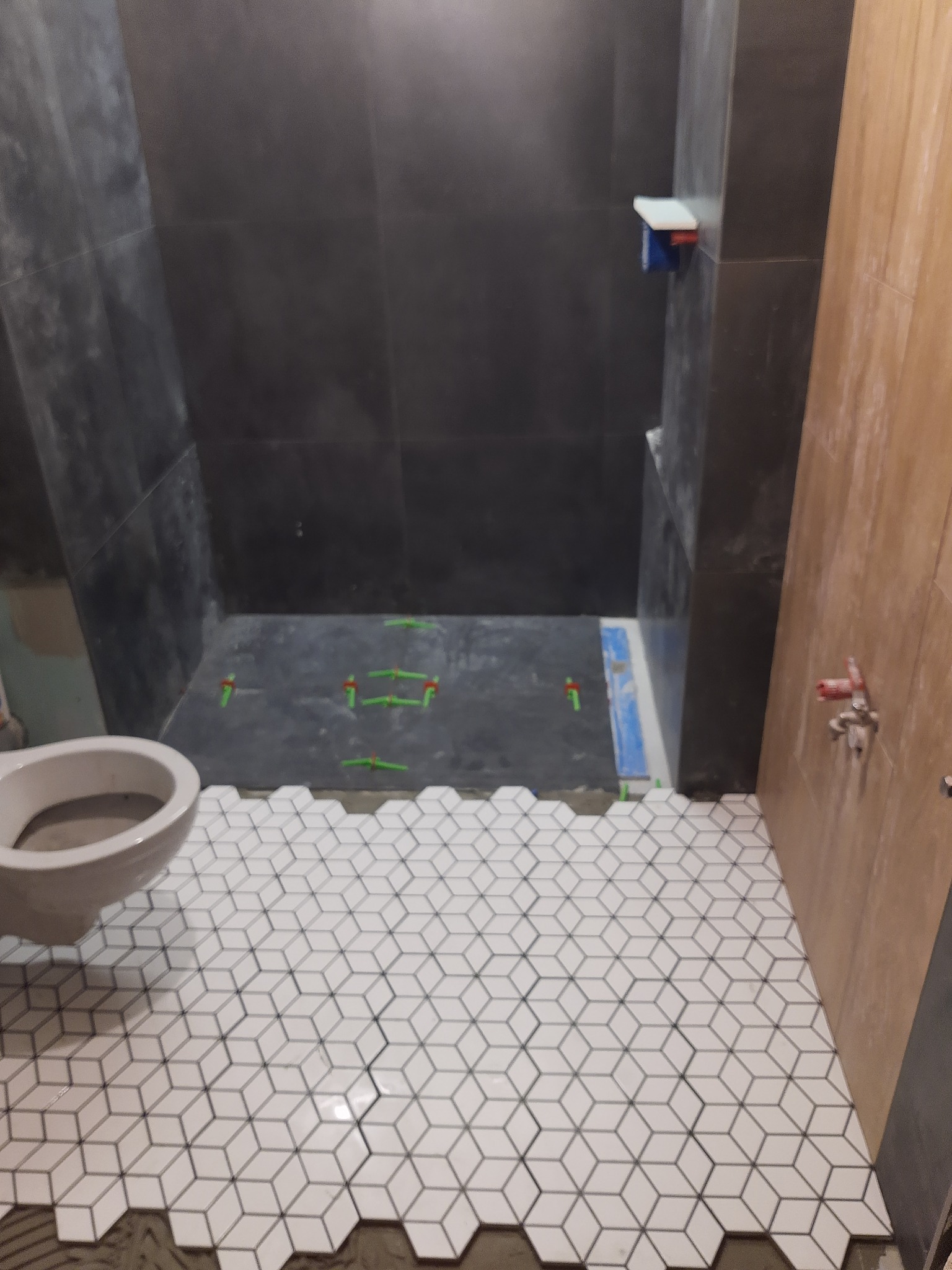 Wnętrze łazienki w trakcie remontu; widoczne czarne płytki ścienne, biała mozaika podłogowa w geometryczny wzór, częściowo ułożona, oraz toaleta bez spłuczki.