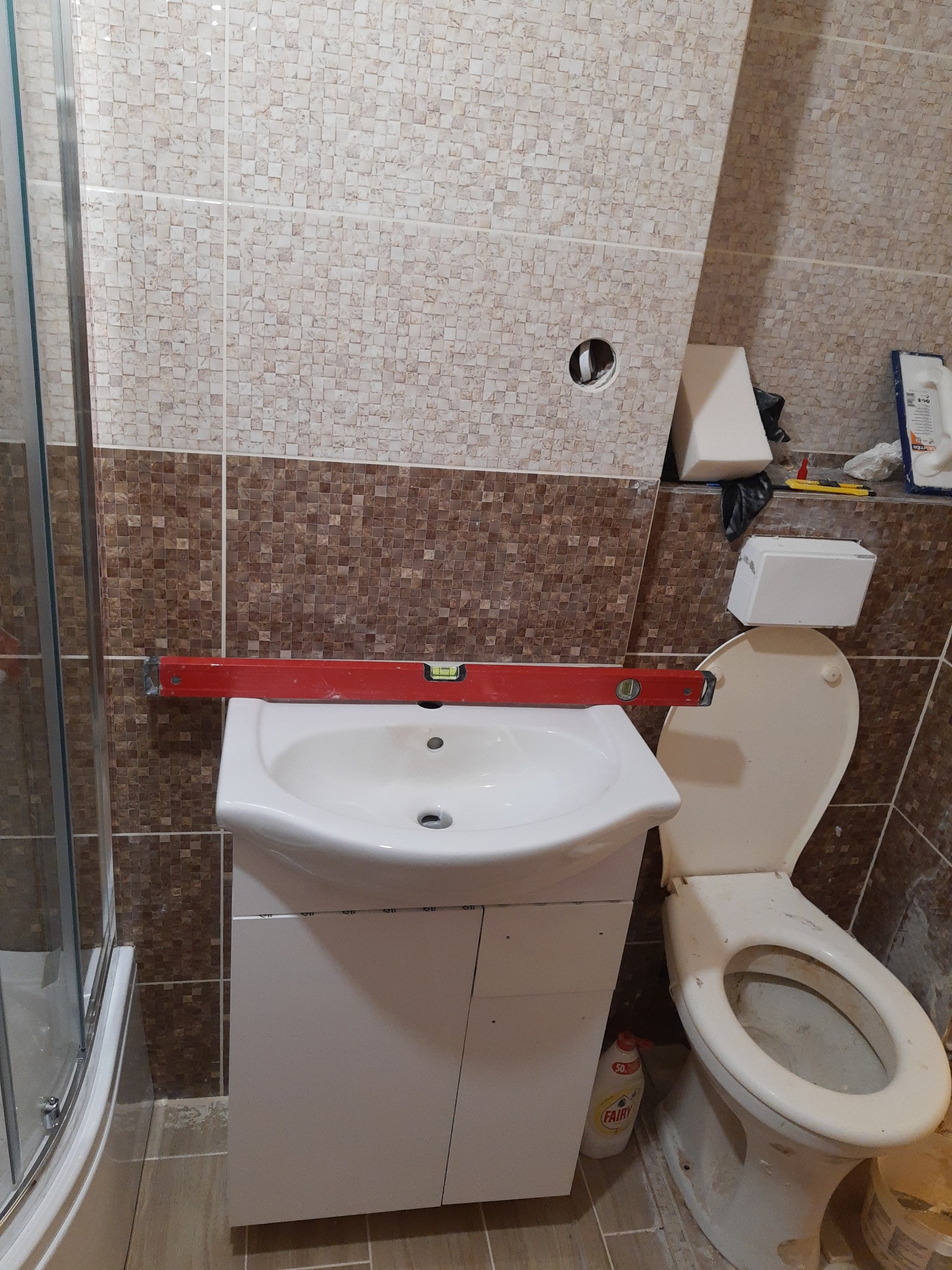 Ustawianie poziomu umywalki z szafką w łazience z mozaikową glazurą, widoczna toaleta i butelka płynu do mycia.