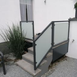 Luk.steel Łukasz Bogdański zpuh - Schody wejściowe z szarą, szklaną balustradą i czarną ramą, otoczone roślinnością i szarym żwirem, z widocznym krzesłem ogrodowym po lewej stronie.