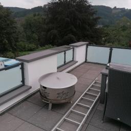 Luk.steel Łukasz Bogdański zpuh - Balkon z szarymi płytkami, otoczony niskimi ściankami z mlecznymi szklanymi panelami, na tle zalesionych wzgórz. Na balkonie znajduje się metalowy grill z ażurowym wzorem, aluminiowa drabina oparta...