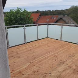 Luk.steel Łukasz Bogdański zpuh - Balkon z drewnianą podłogą i nowoczesną balustradą ze szkła matowego i metalowych słupków, widok na okoliczne domy i zieleń.