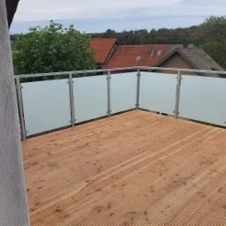 Balkon z drewnianą podłogą i nowoczesną balustradą ze szkła matowego i metalowych słupków, widok na okoliczne domy i zieleń.