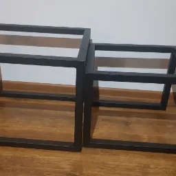 Dwie metalowe ramy w kształcie prostopadłościanów o kwadratowej podstawie, pomalowane na czarno, ułożone jedna w drugiej na drewnianej podłodze, widoczne otwory montażowe.