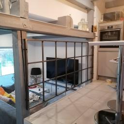 Luk.steel Łukasz Bogdański zpuh - Metalowa konstrukcja z kwadratowymi podziałami oddzielająca część kuchenną od salonu, widok na sofę z kolorowymi poduszkami i telewizor w tle.