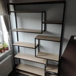 Regał metalowy z drewnianymi półkami w stylu industrialnym, ustawiony przy białej ścianie, widoczne rośliny doniczkowe na parapecie obok okna.