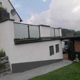 Luk.steel Łukasz Bogdański zpuh - Biały balkon z matowymi szklanymi panelami i ciemnymi ramami, widok z zewnątrz, z roślinami w czarnej donicy na górze, kamienną podmurówką i żwirowym podłożem.