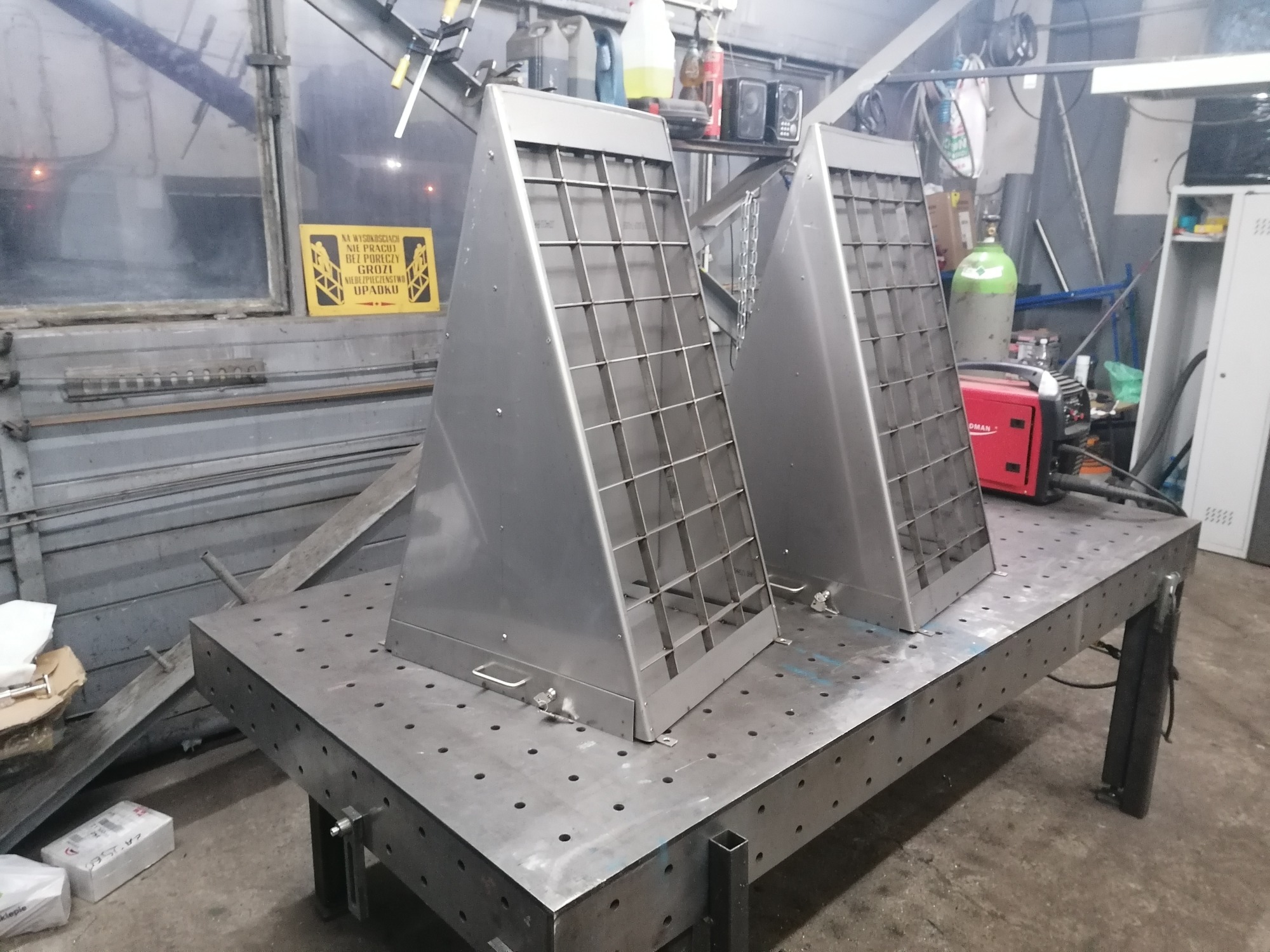 Dwa metalowe, stożkowe filtry z kratką, umieszczone na perforowanym stole spawalniczym w warsztacie, obok butli z gazem i spawarki.