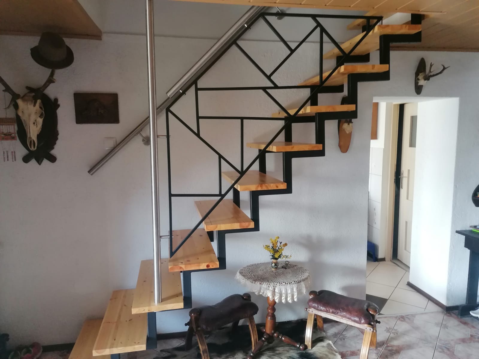 Schody drewniane z czarną, geometryczną konstrukcją metalową i srebrną poręczą, w tle ściana z dekoracjami myśliwskimi i stolik z kwiatami.