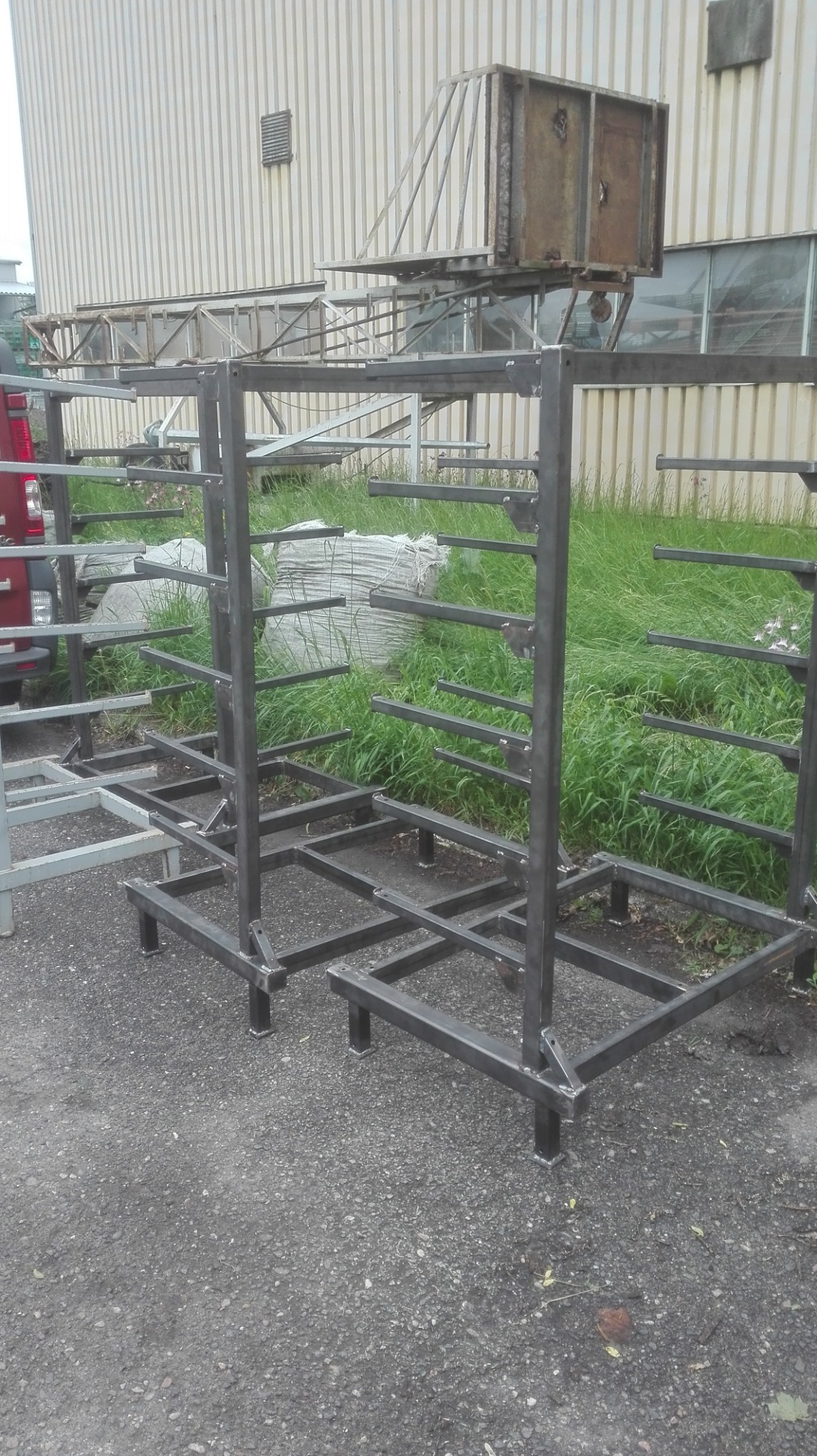 Surowe, metalowe stelaże z poziomymi wspornikami, prawdopodobnie elementy konstrukcyjne do balustrady lub ogrodzenia, stojące na zewnątrz na utwardzonej powierzchni, z trawą i budynkiem w tle.