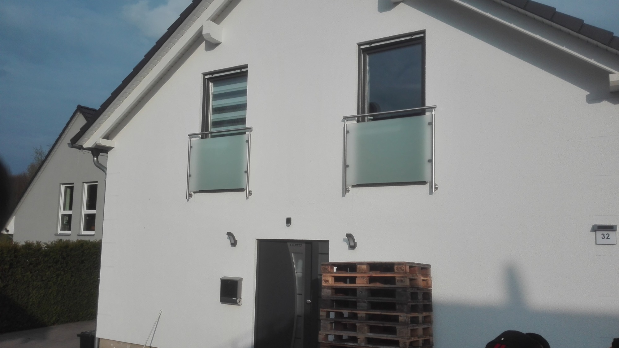 Biały dom z dwoma balkonami zabezpieczonymi balustradami ze stali nierdzewnej i szkła mrożonego, palety drewniane przy wejściu.