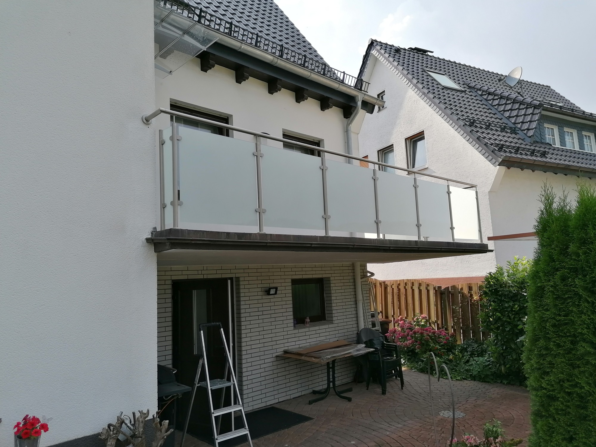 Balkon z matowym szkłem i stalowymi słupkami, widok z dołu na tle domu z czarną dachówką, drabina oparta o ścianę, częściowo widoczne narzędzia na stole, kwiaty w donicach.