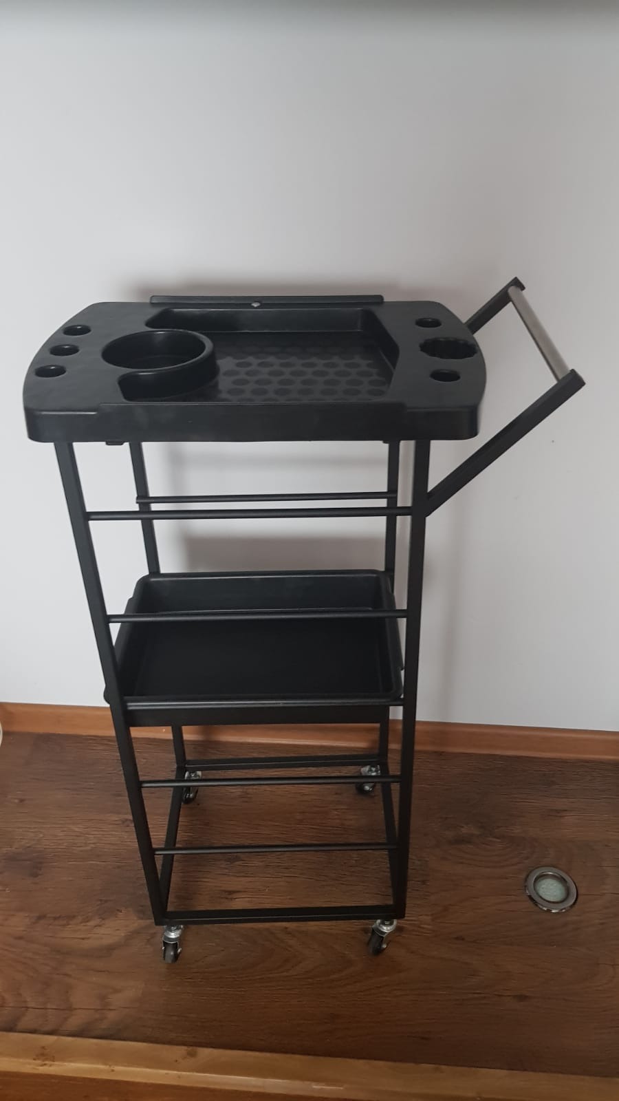 Czarny, metalowy wózek warsztatowy na kółkach z trzema poziomami, widoczny na tle białej ściany i drewnianej podłogi.