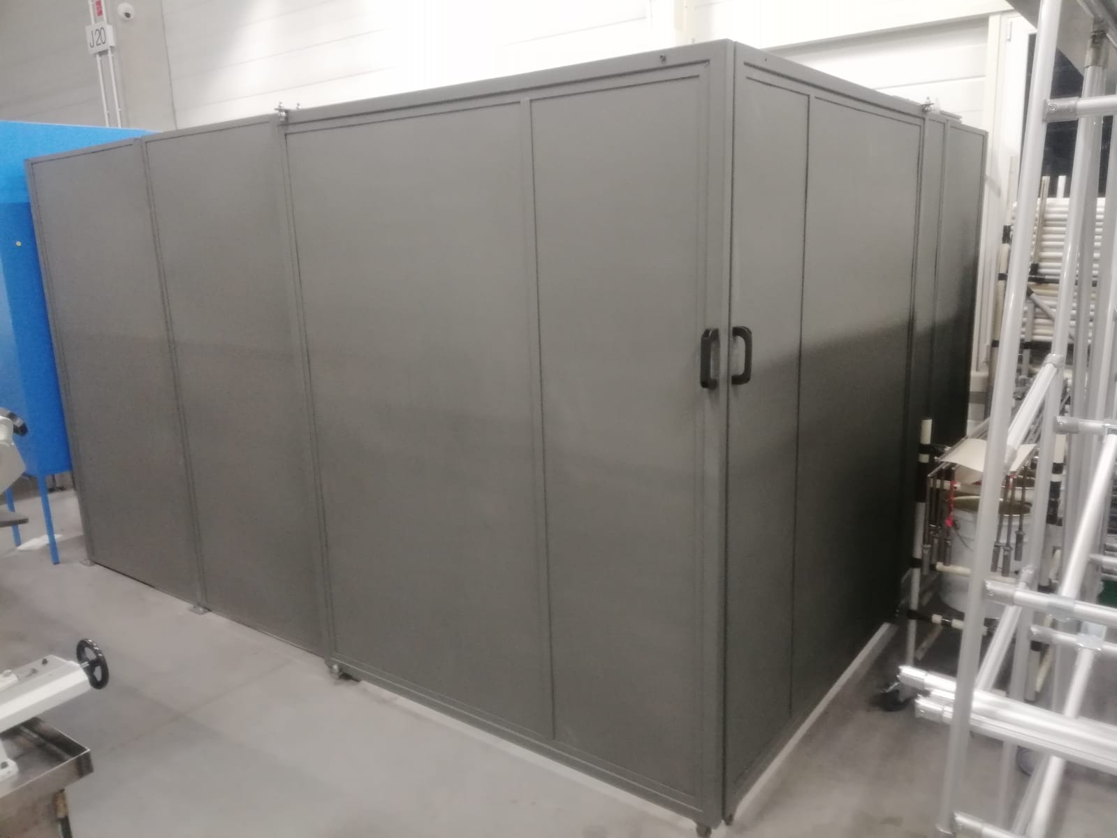 Szary, modułowy system ścianek działowych z drzwiami w hali produkcyjnej, widoczne elementy instalacji i rusztowania.