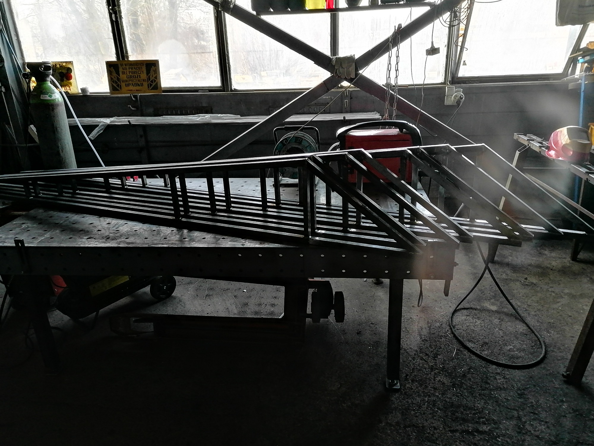 Metalowa konstrukcja kratowa w warsztacie, częściowo zespawana, leżąca na stole spawalniczym, w tle okno i butla z gazem.