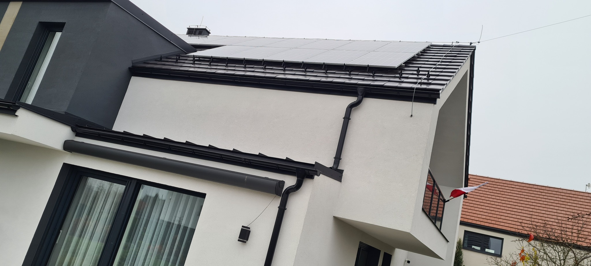 Nowoczesny dom jednorodzinny z panelami fotowoltaicznymi na dachu pokrytym czarną dachówką, widoczne czarne rynny, biała elewacja i okna z czarnymi ramami, na balkonie powiewa polska flaga.