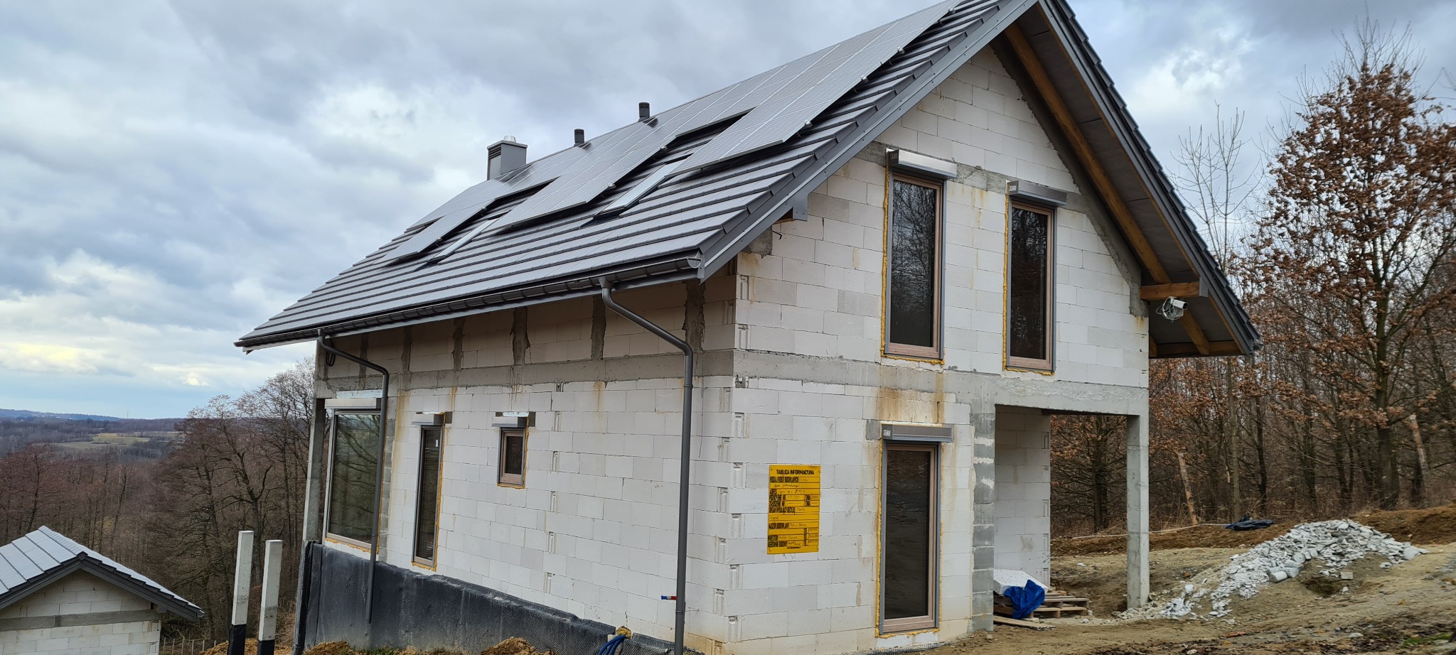 Niedokończony dom z białego bloczka z szarym dachem, na którym zamontowano panele słoneczne. Widoczne orynnowanie i okna z brązowymi ramami. Na ścianie wisi żółta tablica informacyjna. W tle las...