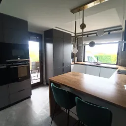 Nowoczesna kuchnia z szarymi szafkami, drewnianym blatem, zielonymi krzesłami i designerską lampą wiszącą, widok na balkon i zieleń za oknem.