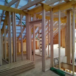 Wnętrze w trakcie budowy domu, widoczne drewniane szkielety ścian, podłoga z płyt OSB, żółta folia zabezpieczająca, drabina i narzędzia w tle.