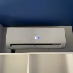 Biały klimatyzator Haier zamontowany na szarej ścianie pod niebieskim sufitem, widoczny wyświetlacz z temperaturą 17 stopni, poniżej białe półki.