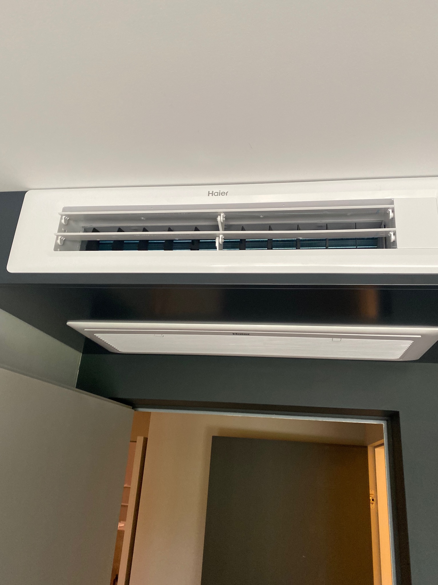 Biały klimatyzator kanałowy Haier zamontowany pod sufitem, widoczne lamele wylotowe, fragment ściany i drzwi w tle.