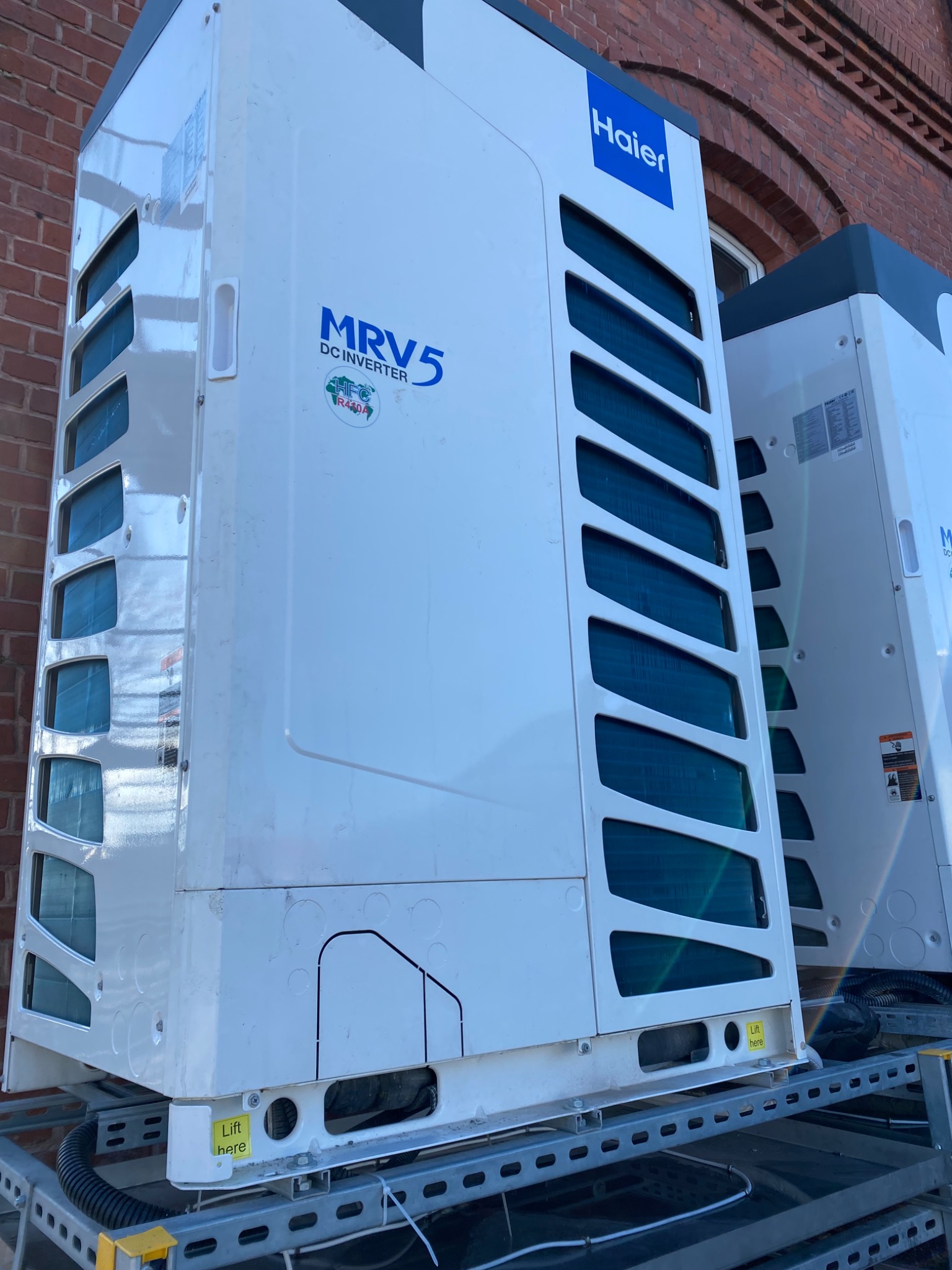 Dwa białe klimatyzatory MRV5 DC Inverter firmy Haier, umieszczone na stalowej konstrukcji na tle ceglanego budynku. Widoczne otwory wentylacyjne, naklejki informacyjne i żółte etykiety z napisem...