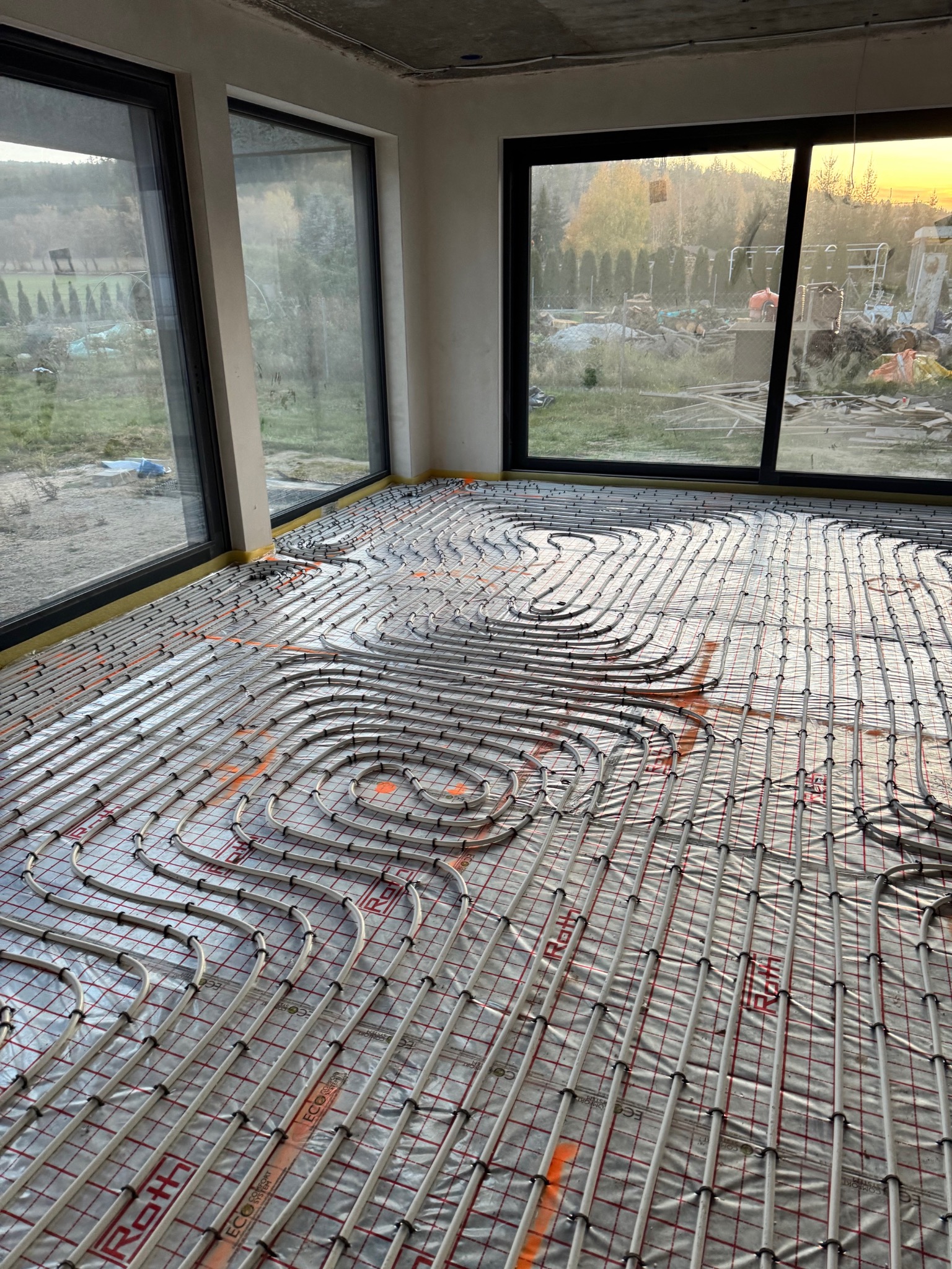 Rozłożone rury ogrzewania podłogowego Roth Eco Comfort System na folii z siatką, widok z góry w surowym wnętrzu z dużymi oknami i widokiem na zewnątrz.