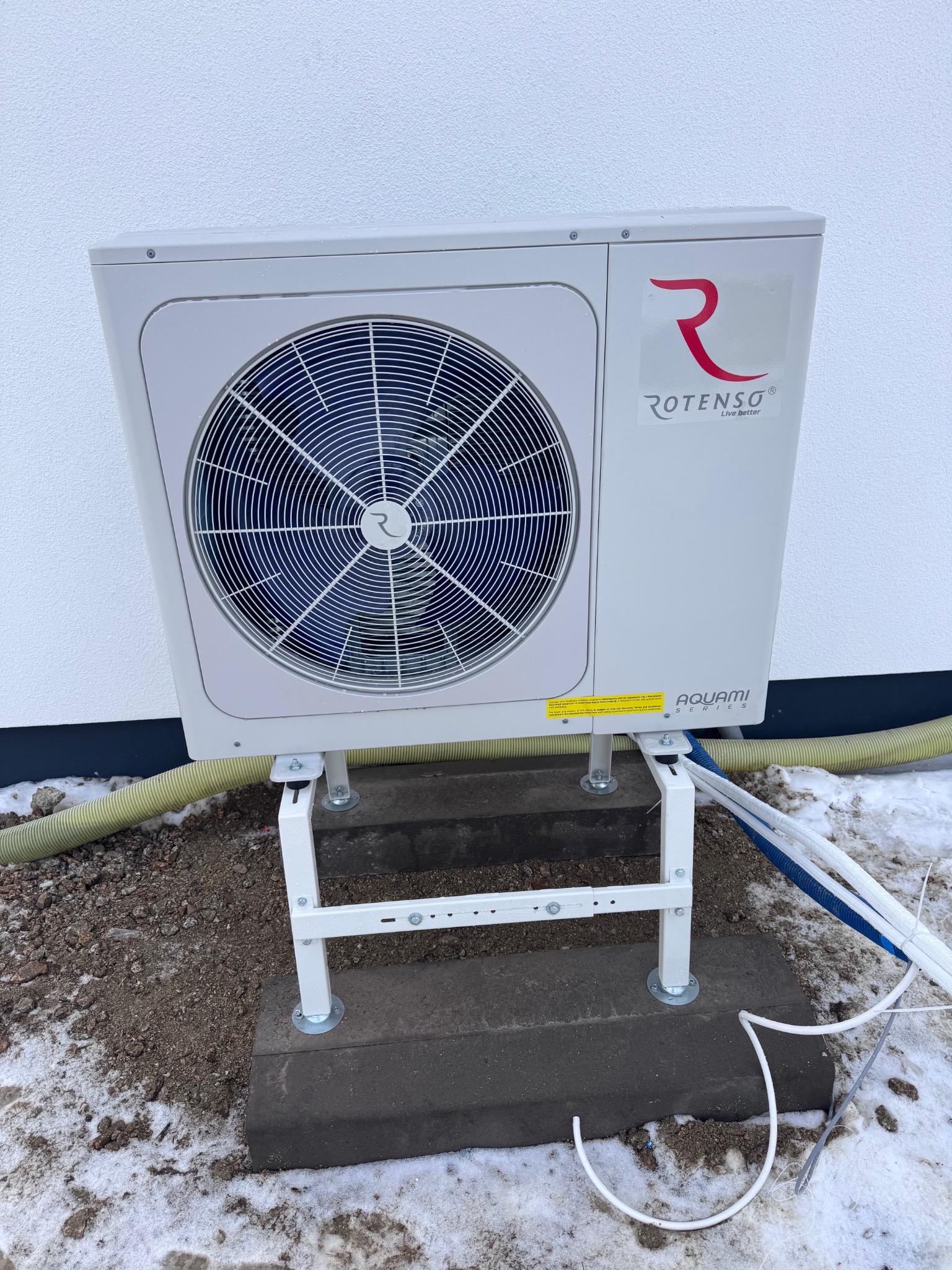 Zewnętrzna jednostka pompy ciepła Rotenso Aquami na białym stelażu, zamontowana przy ścianie budynku. Widoczne przewody i fragment ośnieżonego gruntu.