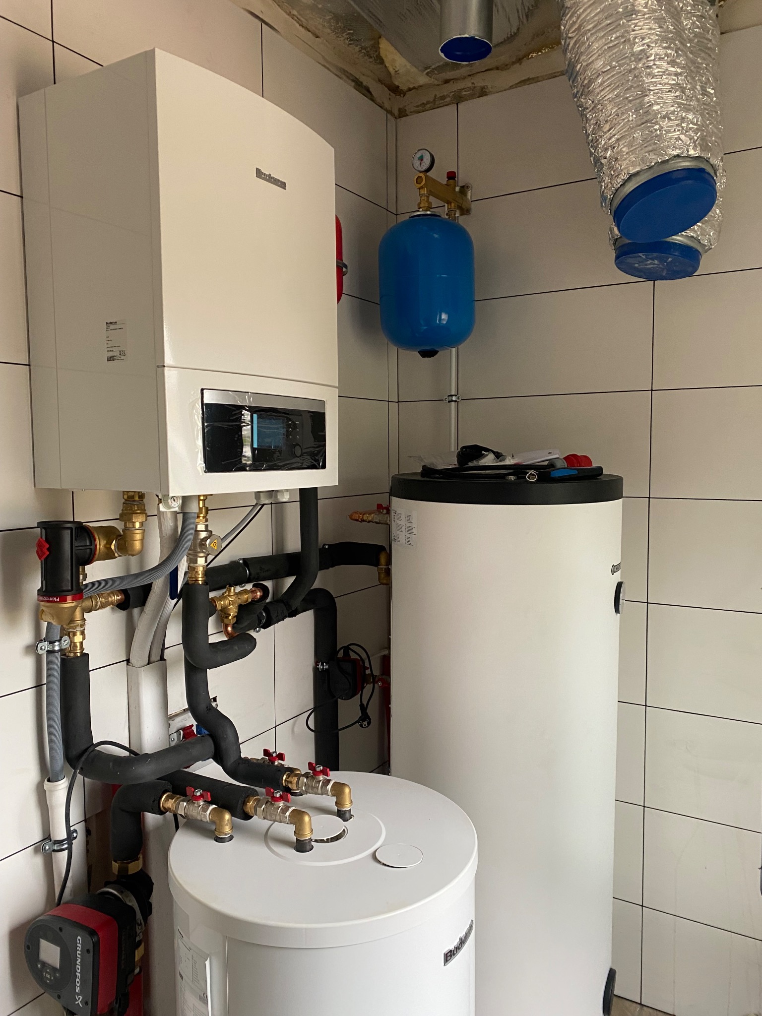 Biały piec gazowy Buderus z podłączonymi rurami i armaturą, obok biały zasobnik ciepłej wody, niebieski zbiornik wyrównawczy i aluminiowy przewód wentylacyjny w pomieszczeniu wyłożonym białymi...