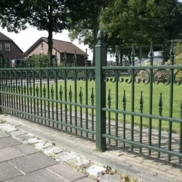 Zielone, metalowe ogrodzenie z dekoracyjnymi elementami w kształcie grotów i kół, widoczne z perspektywy chodnika wyłożonego kostką, z domami i zielenią w tle.