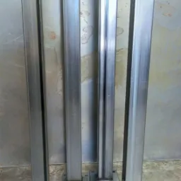 Cztery metalowe słupki o kwadratowym przekroju, przyspawane do kwadratowych podstaw, ustawione na betonowej podłodze z metalową ścianą w tle.