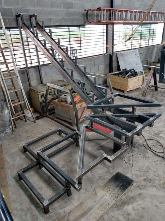 Metalowa konstrukcja schodów w trakcie spawania w warsztacie. Widoczne elementy schodów, drabiny i skrzynia z narzędziami. Surowy, industrialny charakter.